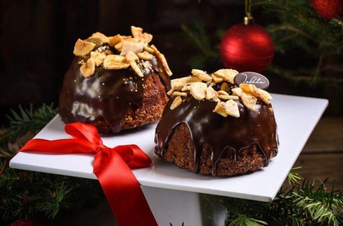 Two Christmas cakes on a white tray. The cakes are covered in chocolate and sprinkled with crushed bananas. On the tray, there's a red ribbon, underneath it pine branches, and red balloons.