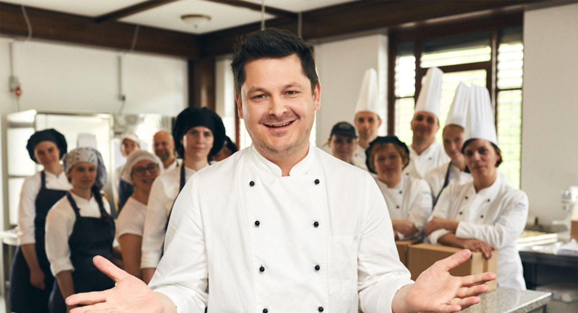 A man in a white chef's uniform, his team in the background with their arms spread wide.