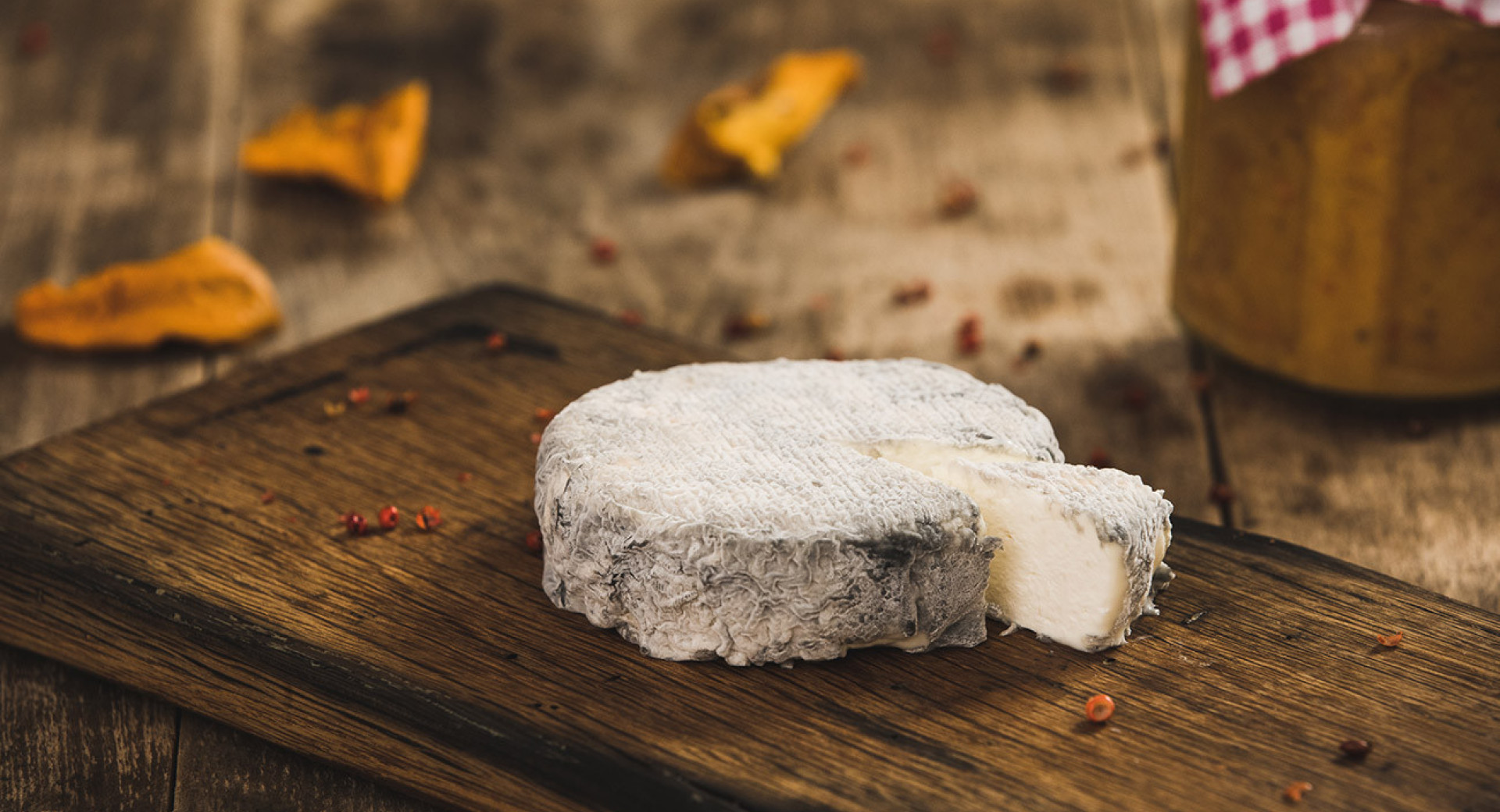 A slice of white cheese with mold on a dark wooden board. In the background, there's a dark table covered with spices and leaves.