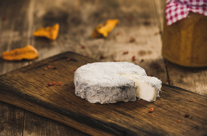 A slice of white cheese with mold on a dark wooden board. In the background, there's a dark table covered with spices and leaves.