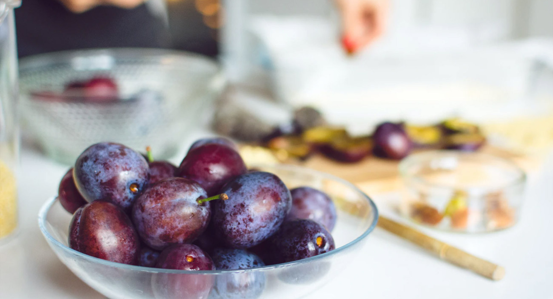 A glass bowl full of plums. In the background, out of focus, is the chef's working surface, with sliced plums on a cutting board.