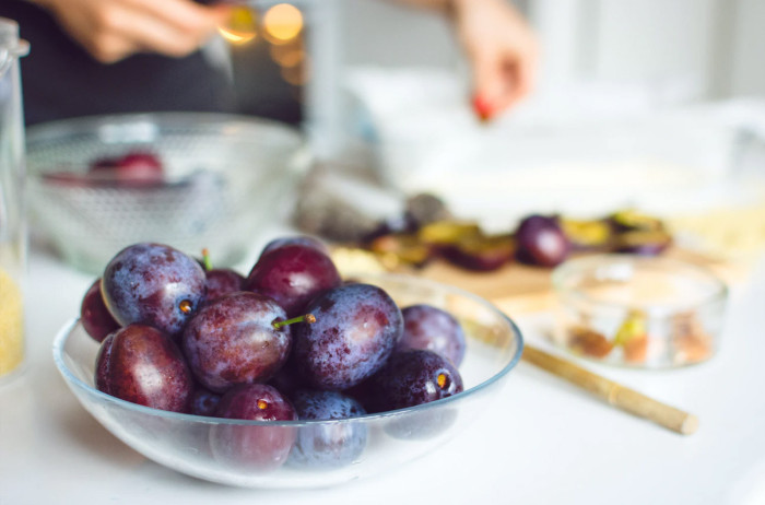 A glass bowl full of plums. In the background, out of focus, is the chef's working surface, with sliced plums on a cutting board.