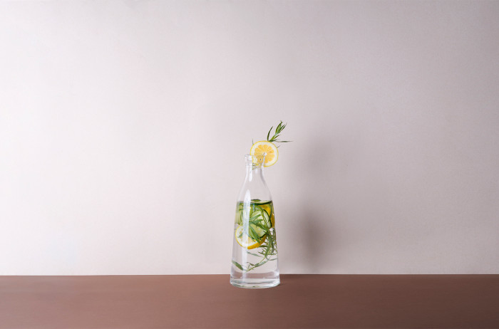  A glass jar on a brown table, with a white background. Inside the jar, there's lemonade with sprigs of mint and slices of lemon.