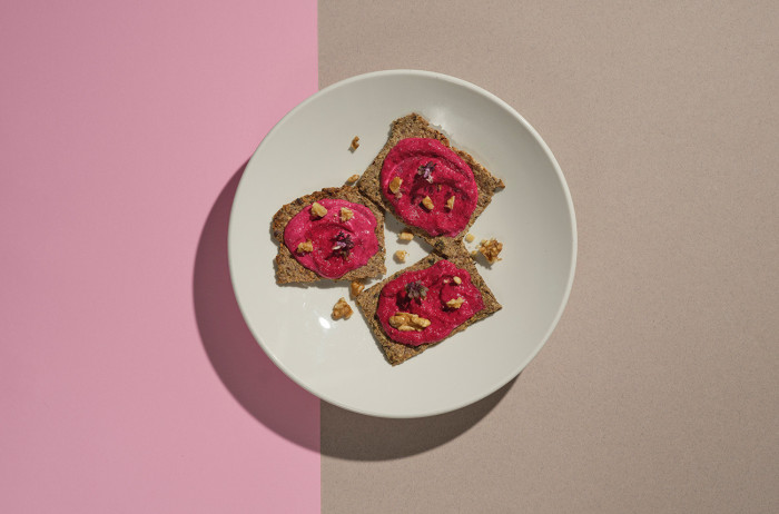 A white plate on a pink and brown background. Neatly arranged brown crackers with pink sauce are placed on the plate. Pumpkin seeds are sprinkled on top for decoration.