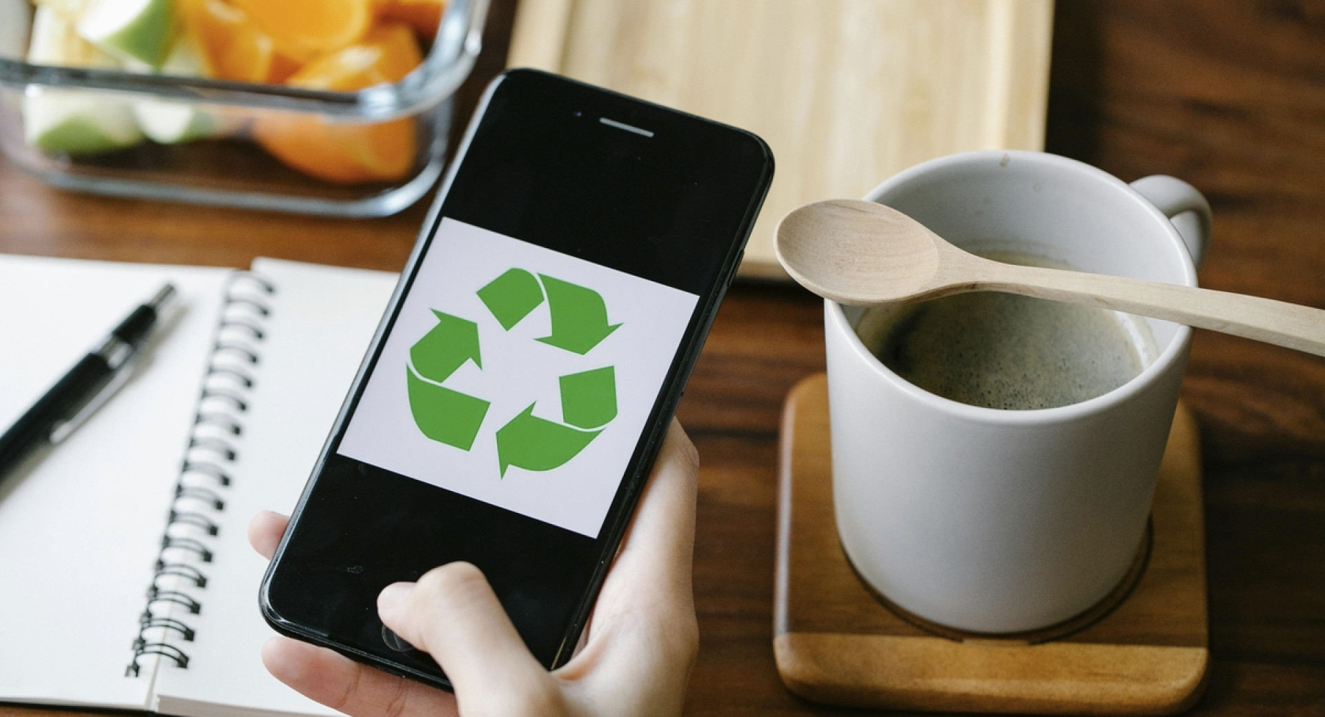 Mobilni telefon v roki z zelenim znakom krožnega gospodarstva, ob njem skodelica in odprt zvezek.