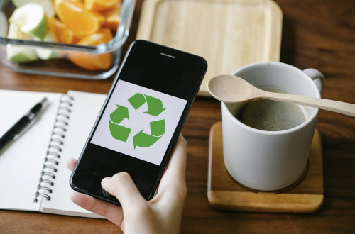Mobilni telefon v roki z zelenim znakom krožnega gospodarstva, ob njem skodelica in odprt zvezek.
