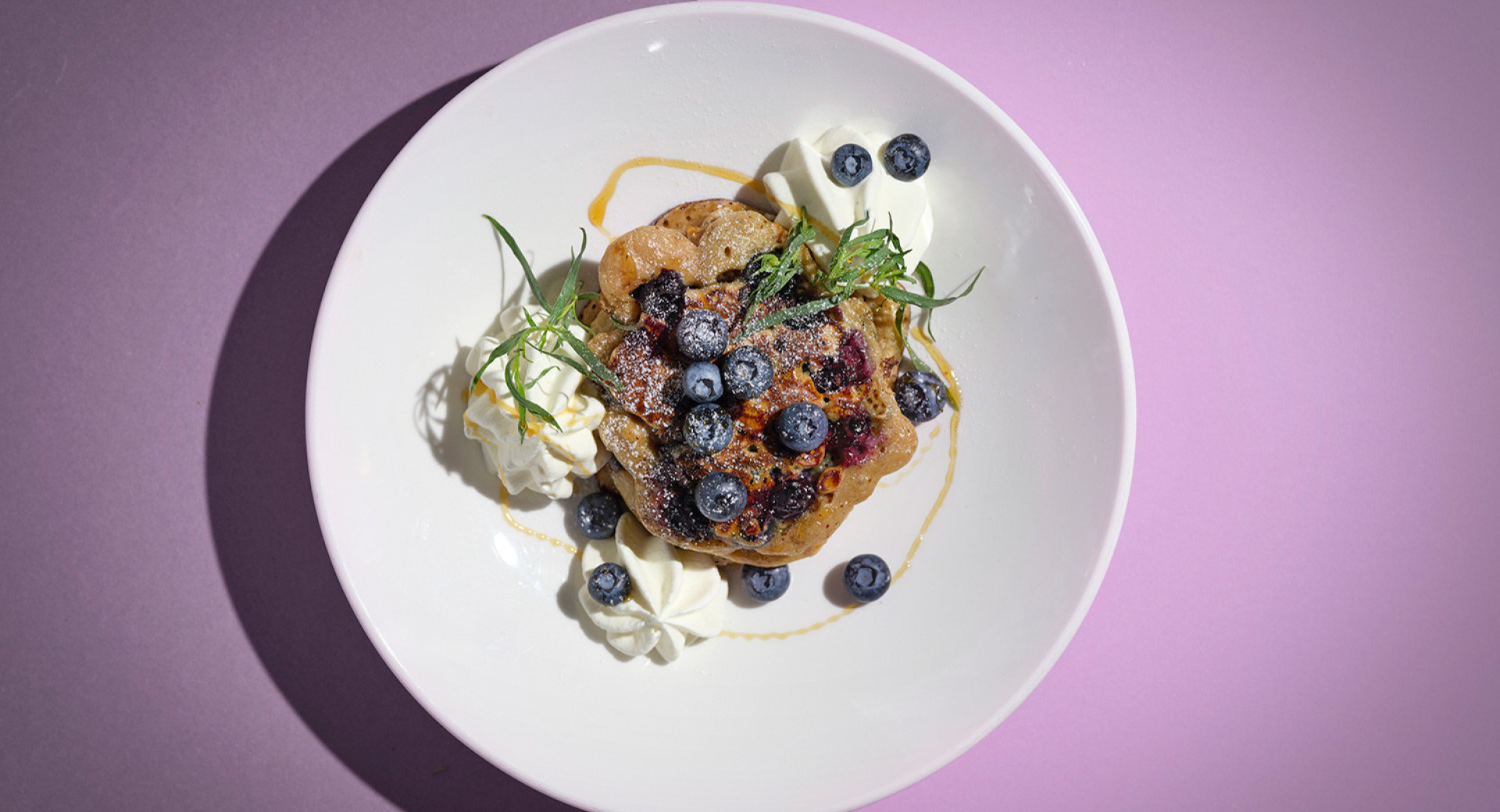 A white plate on a purple and pink base. On the plate, there is a pancake decorated with blueberries, dollops of cream, and sprigs of parsley.