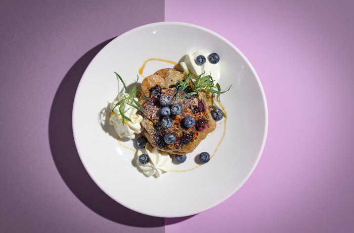 A white plate on a purple and pink base. On the plate, there is a pancake decorated with blueberries, dollops of cream, and sprigs of parsley.