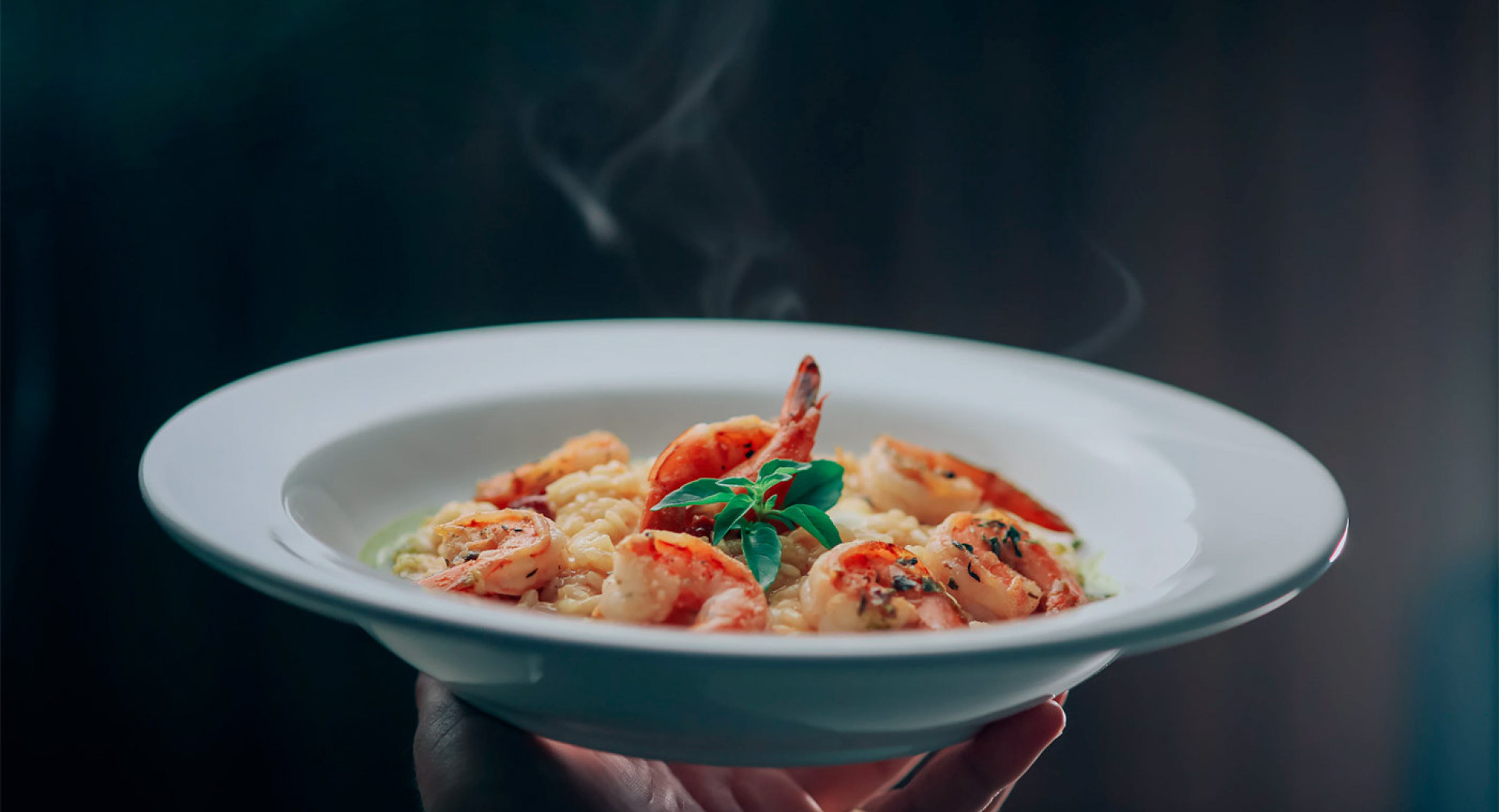 A waiter holds a plate in his hand. On the plate, there is risotto with shrimp neatly arranged on top. Smoke is rising from the plate. The background is dark.