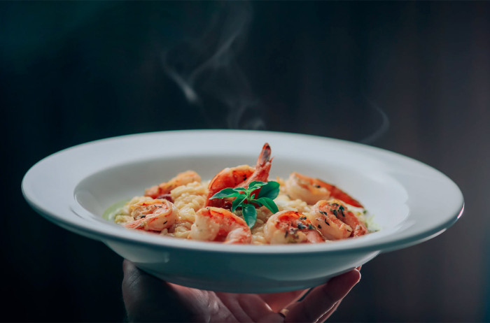 A waiter holds a plate in his hand. On the plate, there is risotto with shrimp neatly arranged on top. Smoke is rising from the plate. The background is dark.