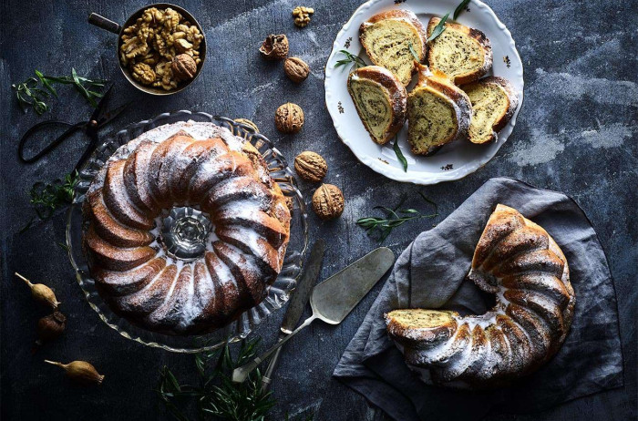 A black background with three plates: one with a whole potica, another with a third missing, and the third with slices. They are dusted with powdered sugar, and there are whole walnuts next to them along with utensils.