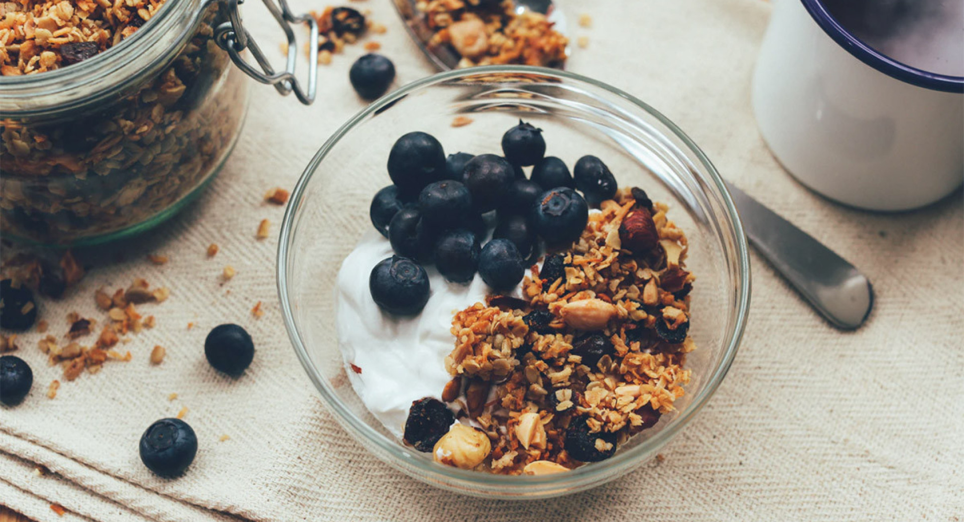 Granola in a glass jar, served with yogurt and blueberries. The jar is placed on a prepared table, with scattered cereal flakes and blueberries around.