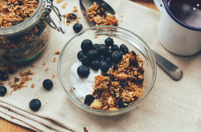 Granola in a glass jar, served with yogurt and blueberries. The jar is placed on a prepared table, with scattered cereal flakes and blueberries around.