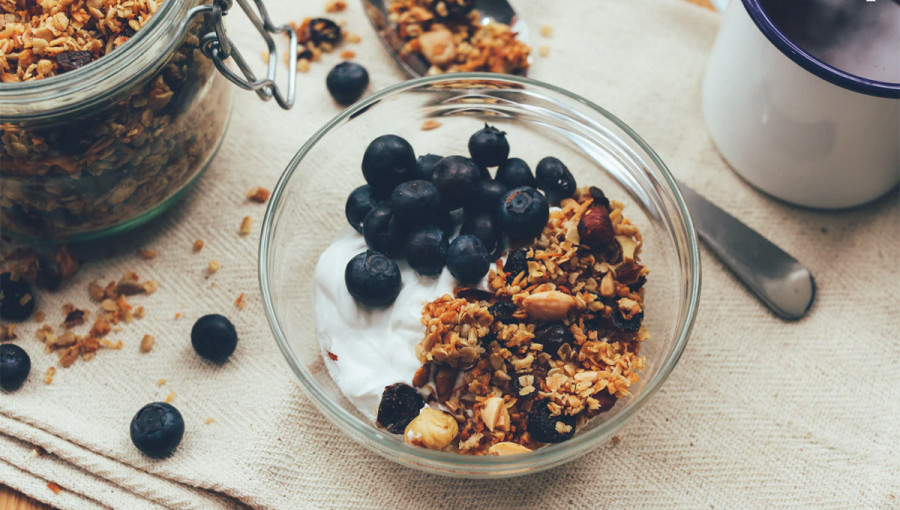 granola v stekleni posodici, postrezena z jogurtom in borovnicami. posoda stoji na pripravljeni mizi, na kateri so raztreseni kosmici in borovnice.