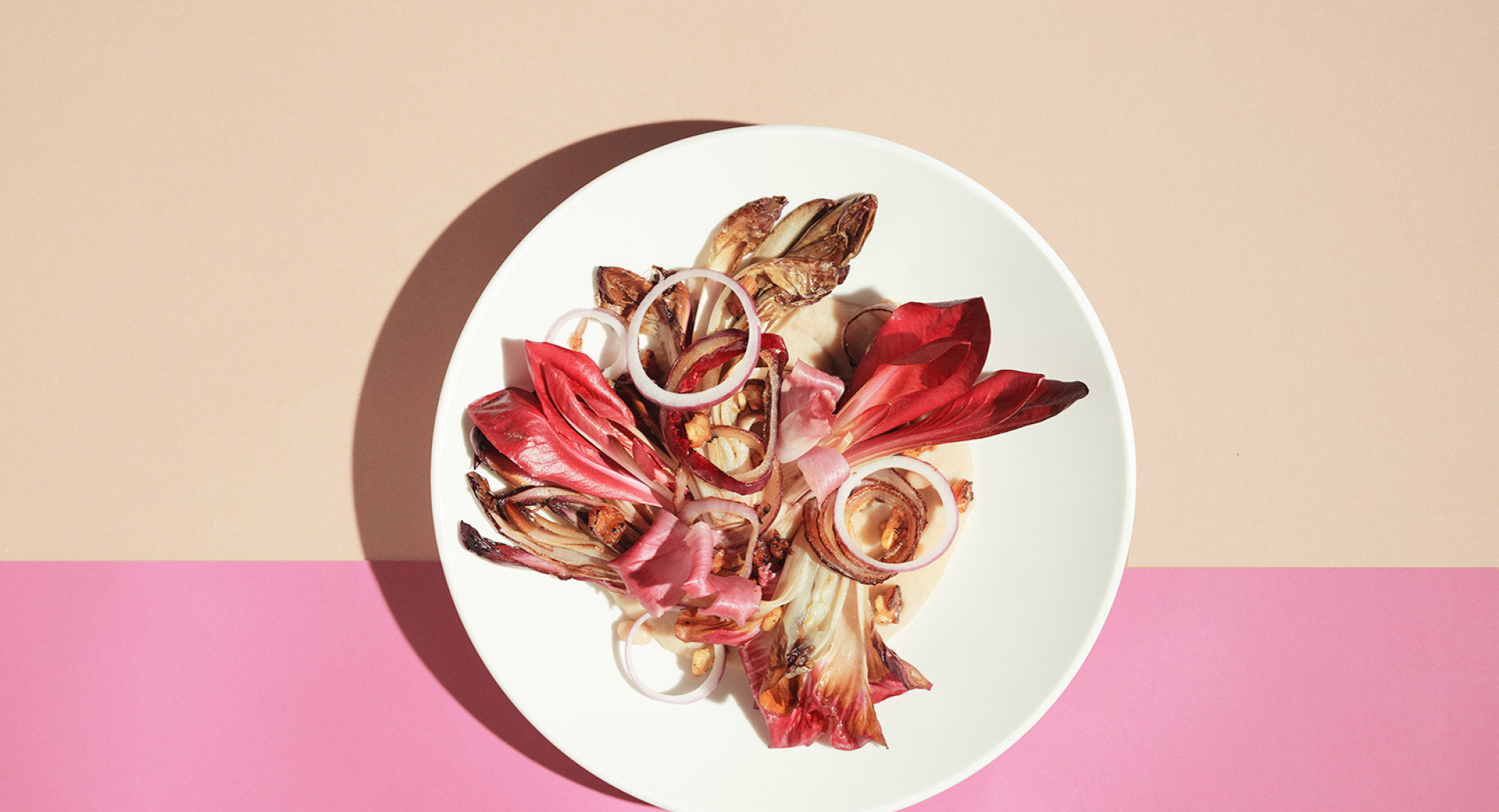  A white plate on a pink and orange background. On the plate, leaves of red radish, sprinkled with onion rings.