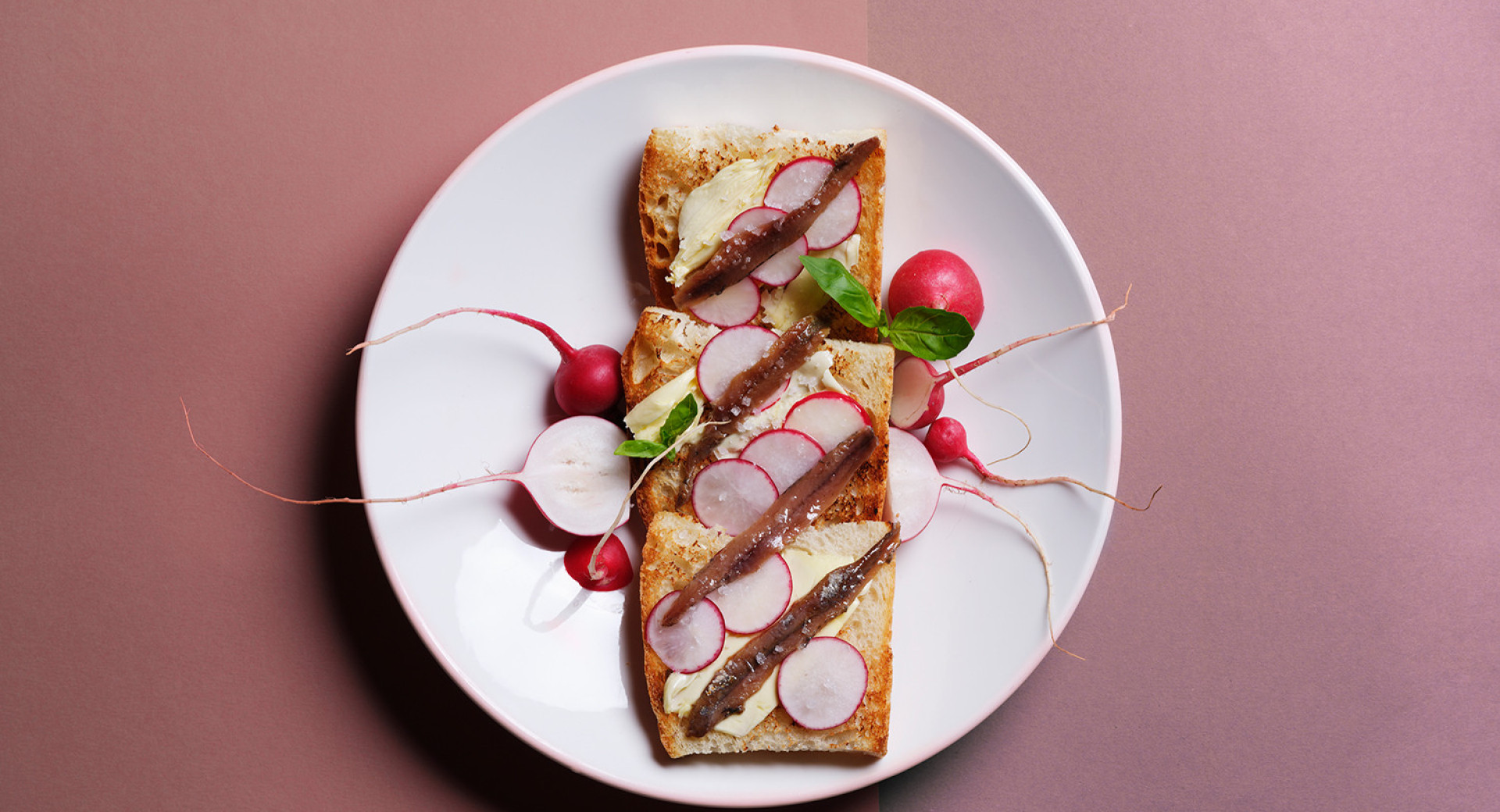 A white plate on a light and dark pink background. On the plate, neatly arranged slices of bread, spread with butter and garnished with radishes and chives.