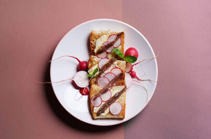 A white plate on a light and dark pink background. On the plate, neatly arranged slices of bread, spread with butter and garnished with radishes and chives.