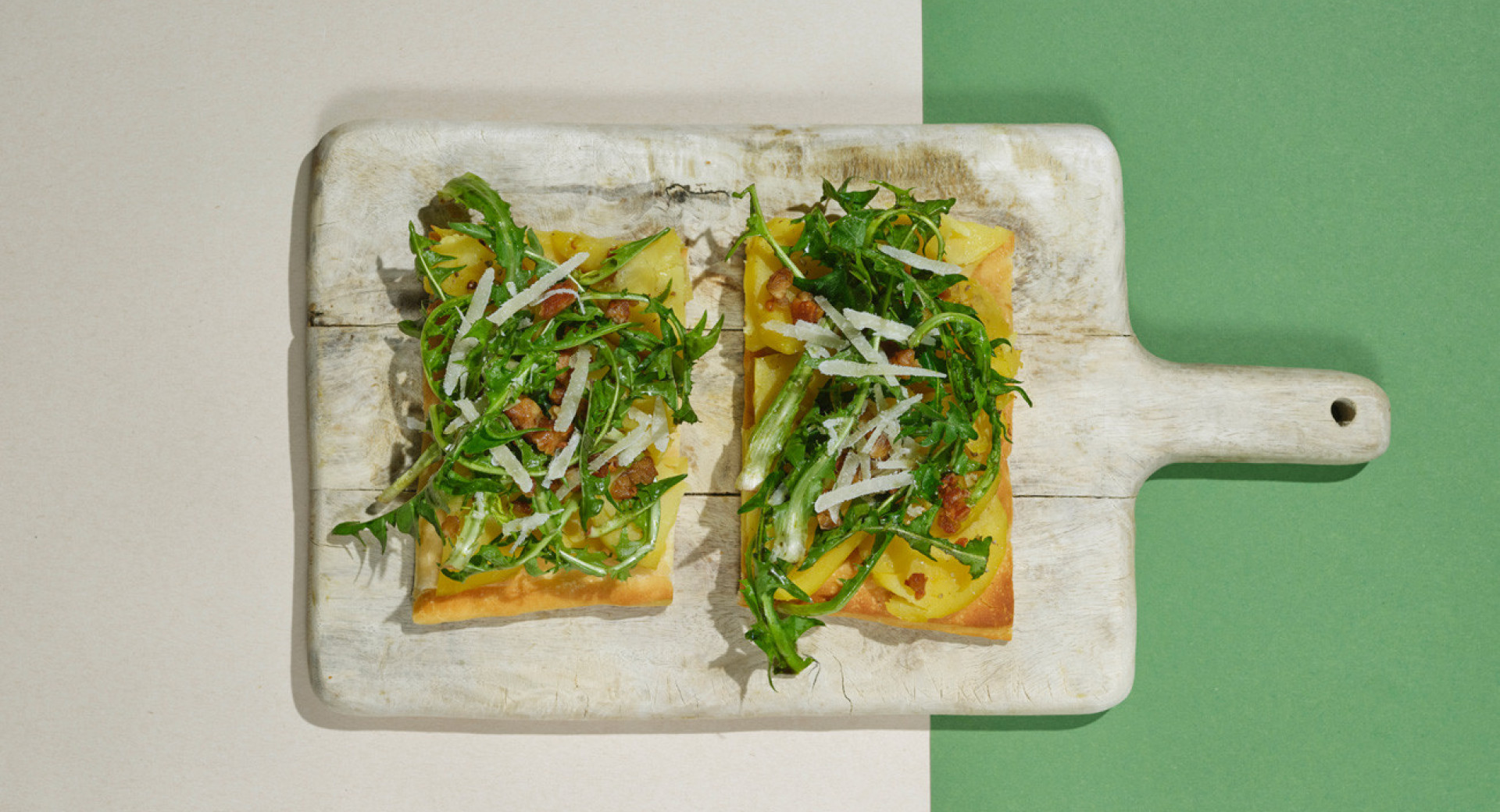 A light wooden board on a beige and green base. On the board, two slices of bread with arugula and grated cheese.