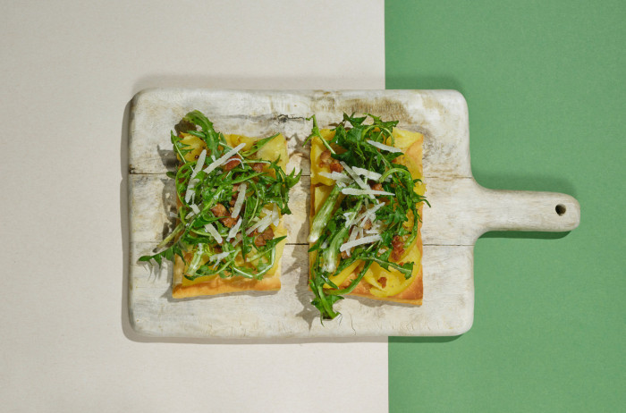 A light wooden board on a beige and green base. On the board, two slices of bread with arugula and grated cheese.