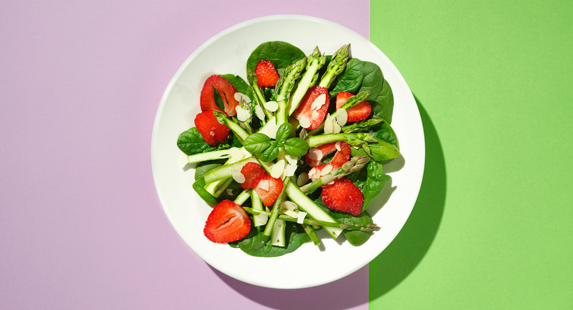 A white plate on a pink and green base. On the plate, neatly arranged salad of asparagus and strawberries.