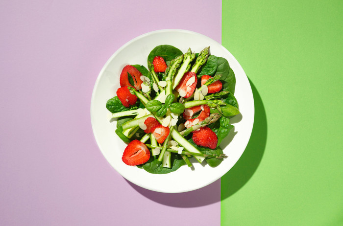 A white plate on a pink and green base. On the plate, neatly arranged salad of asparagus and strawberries.