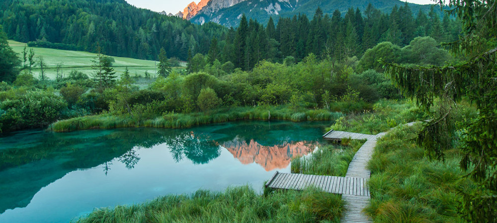 Smaragdno jezerce z leseno potjo med zelenim rastjem in gozdom ter gorami v ozadju ob soncnem zahodu.