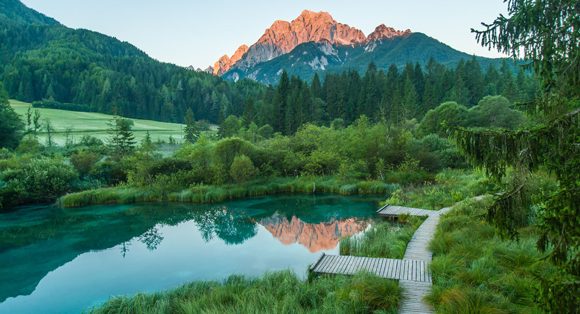 Smaragdno jezerce z leseno potjo med zelenim rastjem in gozdom ter gorami v ozadju ob soncnem zahodu.