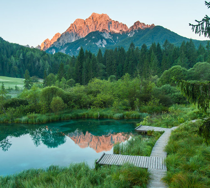Lac emeraude avec un ponton en bois entoure de vegetation verdoyante et de foret, avec des montagnes en arriere-plan au lever du soleil.