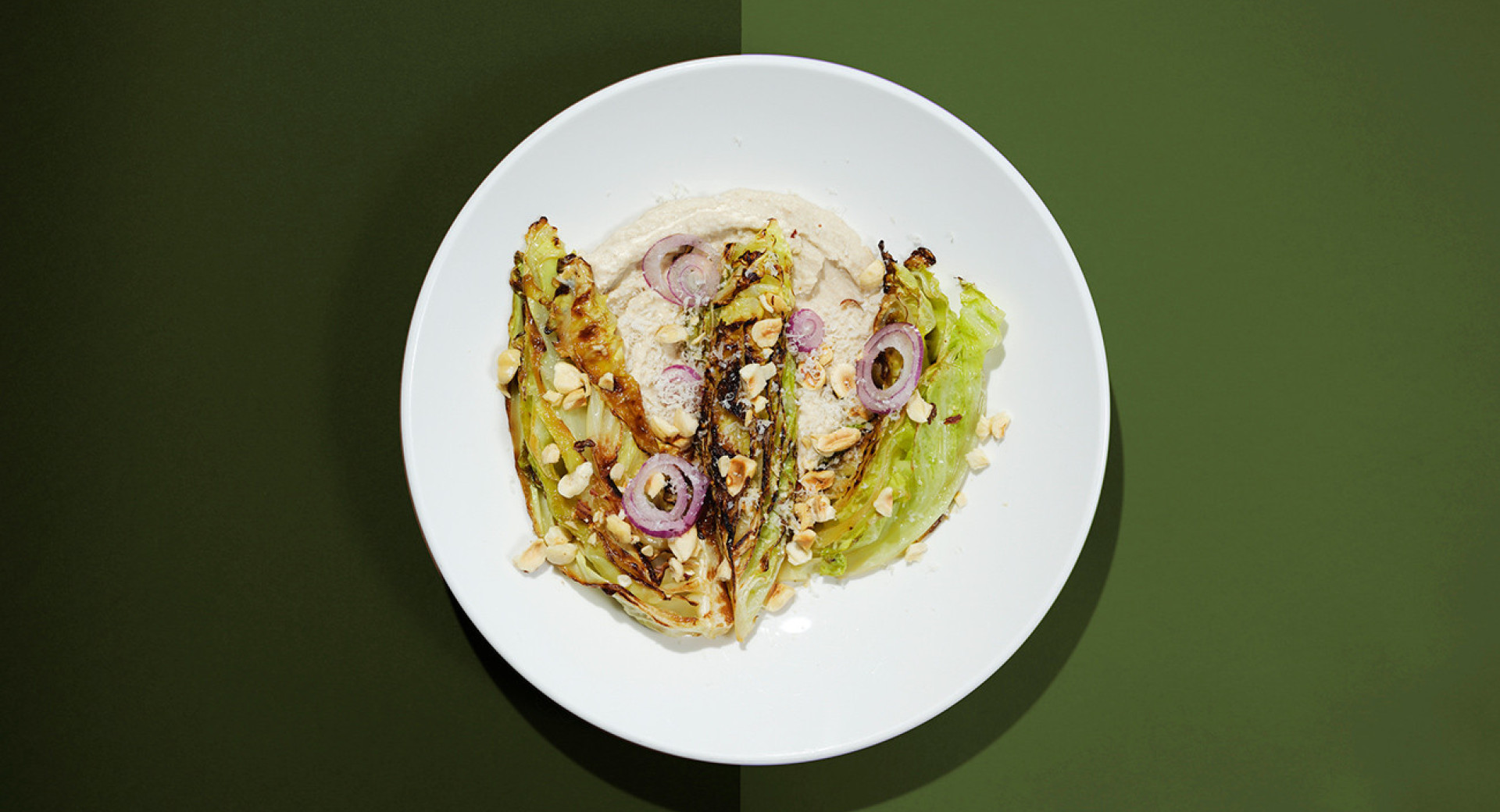 A white plate on a light and dark green base. On the plate, there are roasted cabbage leaves, garnished with slices of onion and sprinkled with hazelnuts.