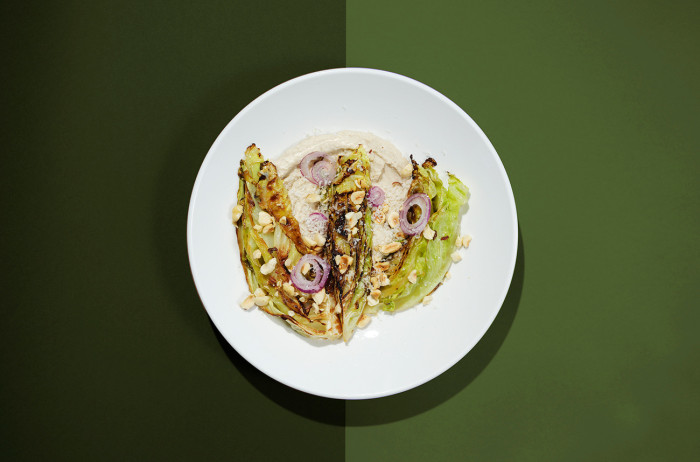 A white plate on a light and dark green base. On the plate, there are roasted cabbage leaves, garnished with slices of onion and sprinkled with hazelnuts.