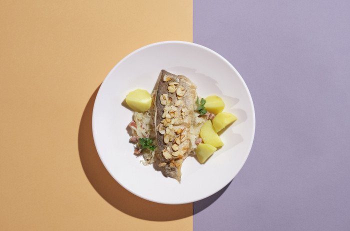 A white plate on an orange and purple base. On the plate, there's a fish fillet and potatoes in pieces, garnished with chopped parsley and almonds.