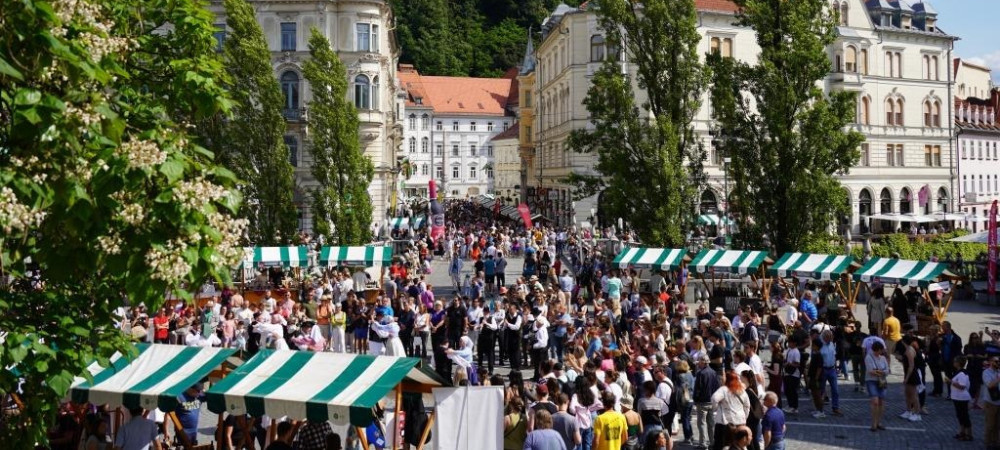 A crowd of people on Preseren trg Square in Ljubljana, gathered around green-and-white stalls, with Ljubljana Castle rising in the background.