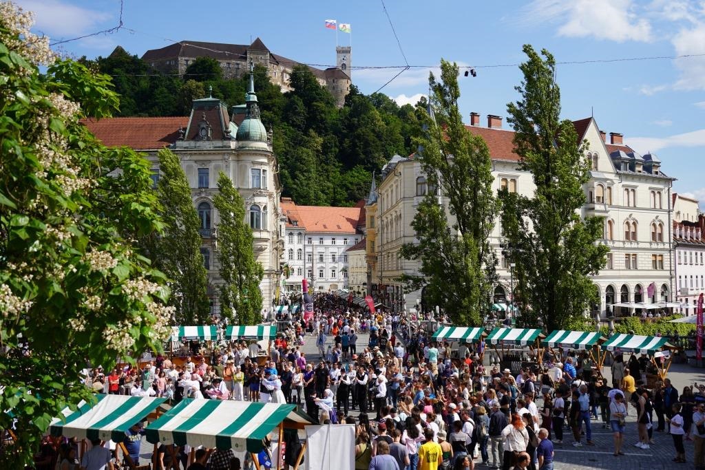 Gruca ljudi na Presernovem trgu v Ljubljani, okoli postavljenih zeleno-belih stojnic, v ozadju pa se dviga Ljubljanski grad.