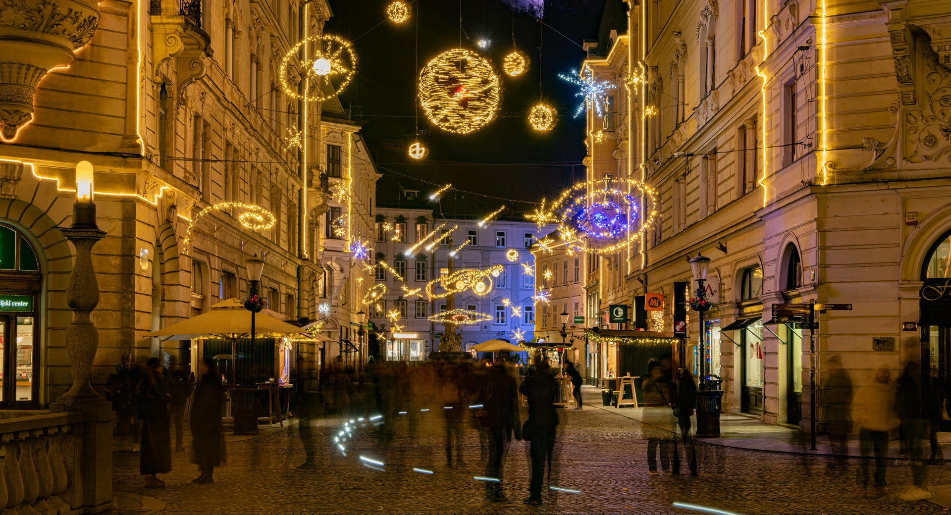 Vista notturna di una strada del centro storico con luci festive, con persone in movimento e un castello illuminato su una collina sullo sfondo.