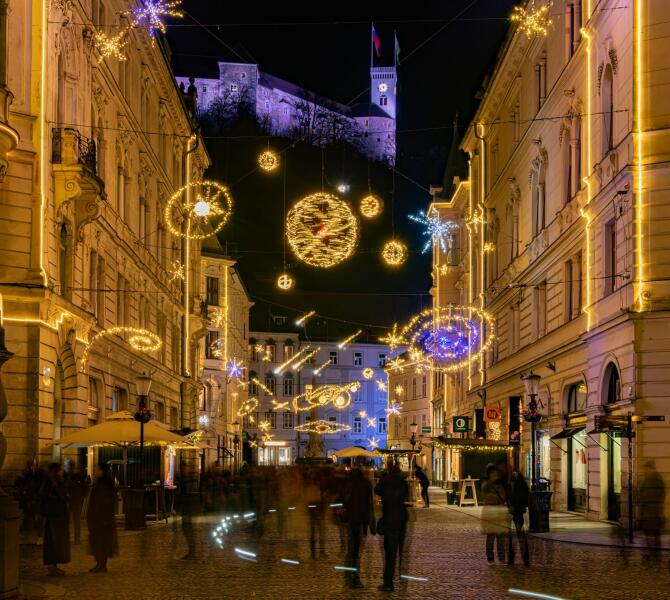 Night view of an old town street with festive lights, with people in motion and an illuminated castle on a hill in the background.