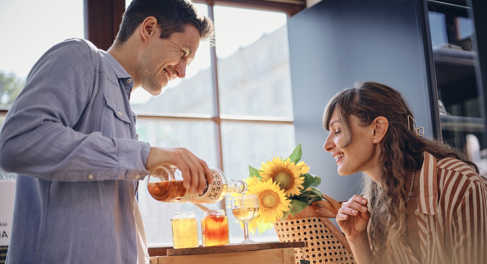 Nasmejan par; moski naliva medico v kozarec in zenska ga opazuje.