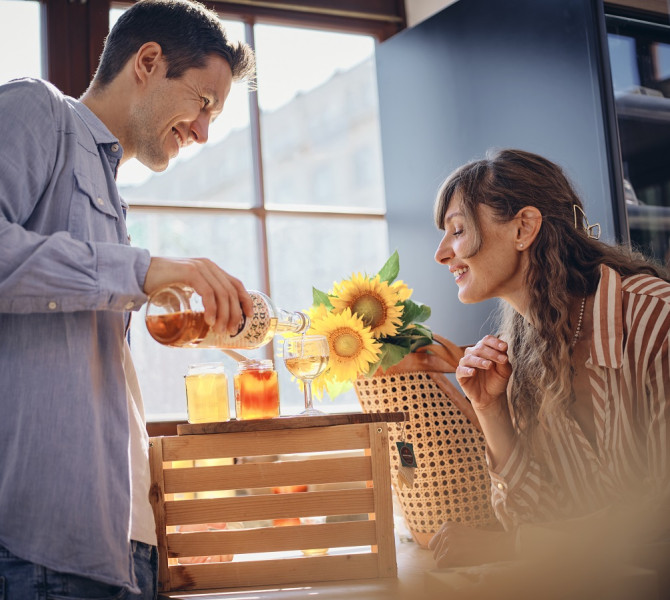 Nasmejan par; moski naliva medico v kozarec in zenska ga opazuje.