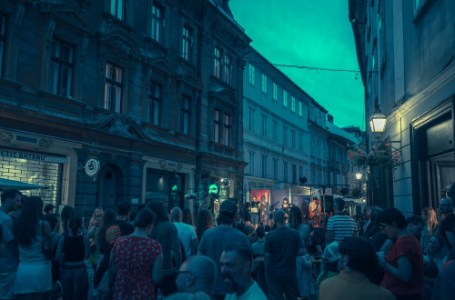Mnozica ljudi na ozki ulici v stari Ljubljani poslusa koncert na malem odru.