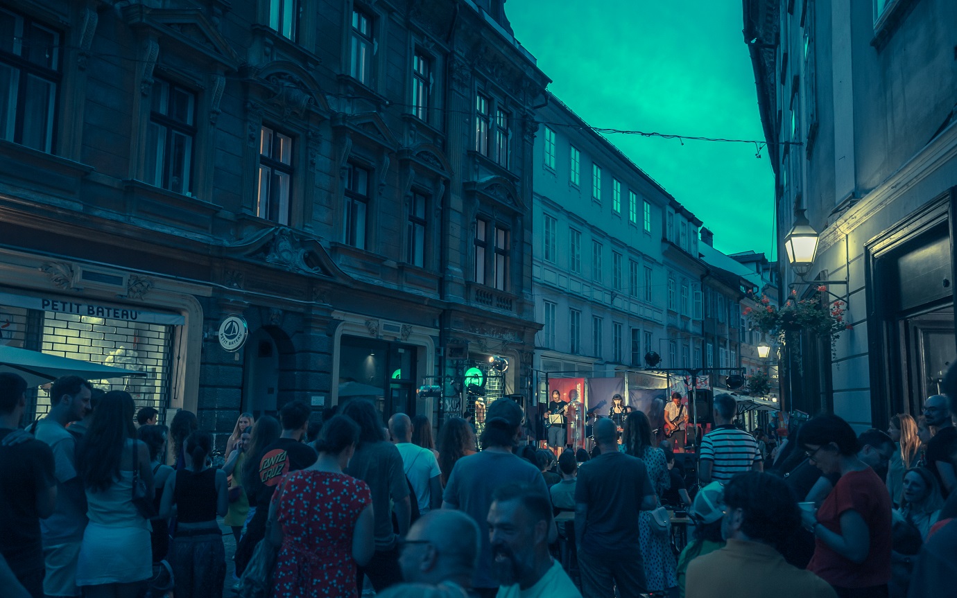 Mnozica ljudi na ozki ulici v stari Ljubljani poslusa koncert na malem odru.