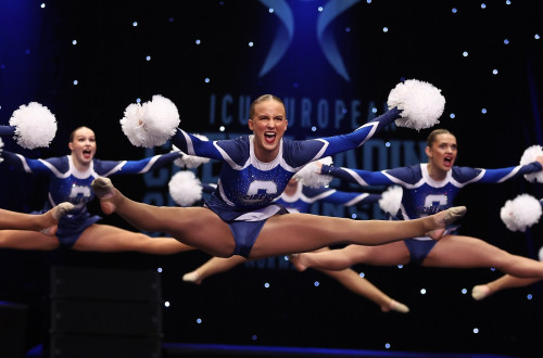 The cheerleaders in blue-and-white uniforms with white pom-poms perform an aerial split jump on stage.