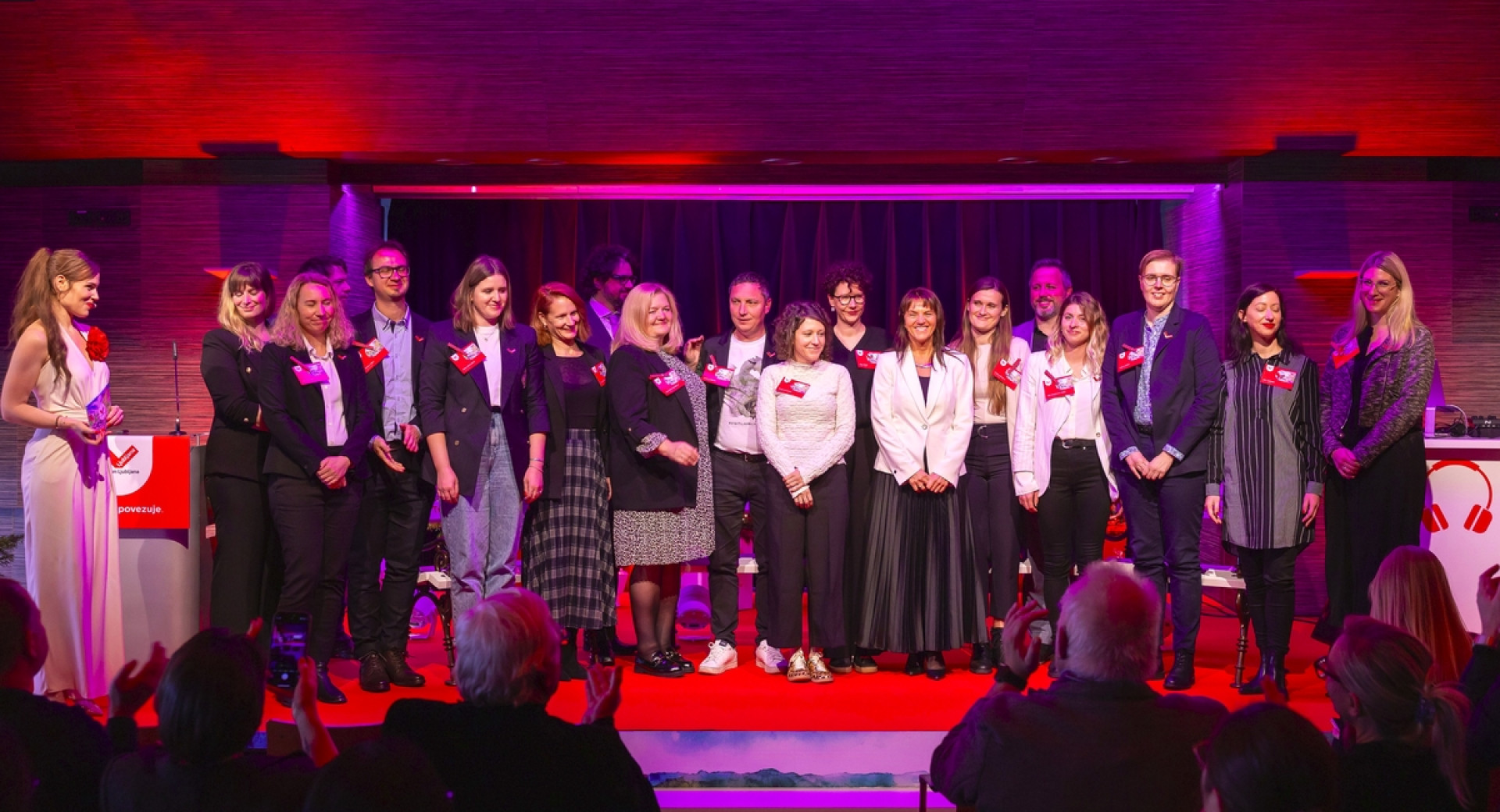 Skupina ljudi stoji na odru, pred njimi občinstvo.