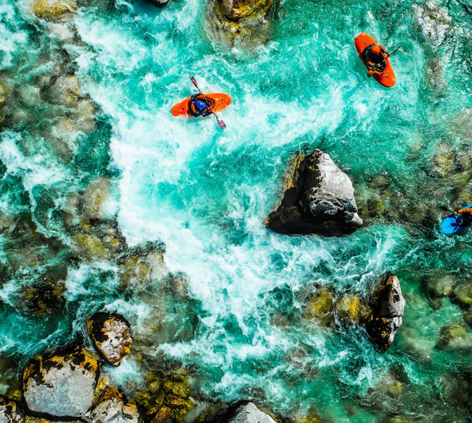 Three kayakers skillfully navigate the rapids of a crystal-clear river among large rocks.