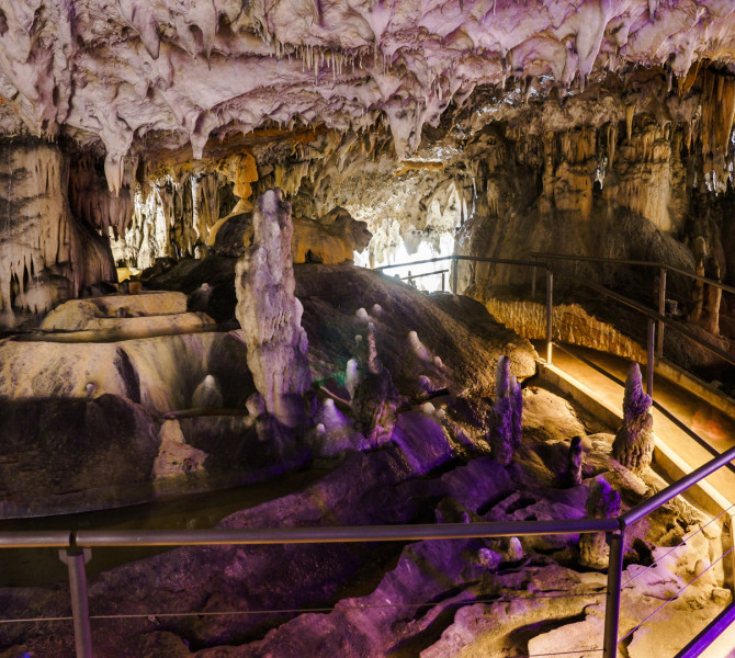 Interno di una grotta con stalattiti e stalagmiti illuminate e un percorso pedonale attrezzato con ringhiera.