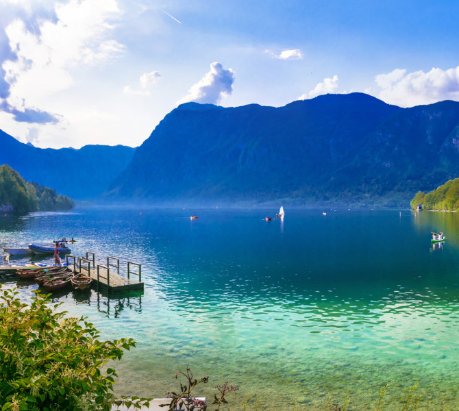 Le acque cristalline del Lago di Bohinj riflettono la rigogliosa foresta circostante e le imponenti Alpi Giulie sotto un cielo luminoso e parzialmente nuvoloso.