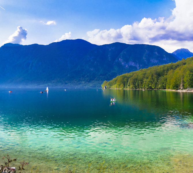 Kristalno cista voda Bohinjskega jezera odseva okoliski bujni gozd in visoke Julijske Alpe pod svetlim, delno oblacnim nebom.