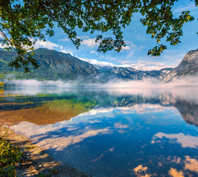 Jutranja meglica se dviga nad mirno povrsino Bohinjskega jezera, ki odseva okoliske gore in drevesa pod svetlo modrim nebom.