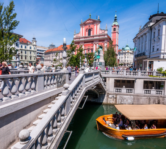 Ein Holzboot fährt unter einer dreifachen Brücke hindurch, im Hintergrund eine rote Kirche.