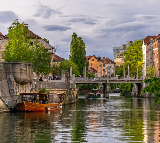 Ein Holzboot an der Anlegestelle am Fluss, im Hintergrund eine Brücke, grüne Ufer und Bürgerhäuser.