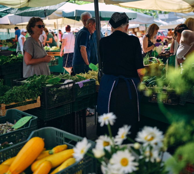 Une dame âgée vendant des légumes au marché, des gens autour de son étal.