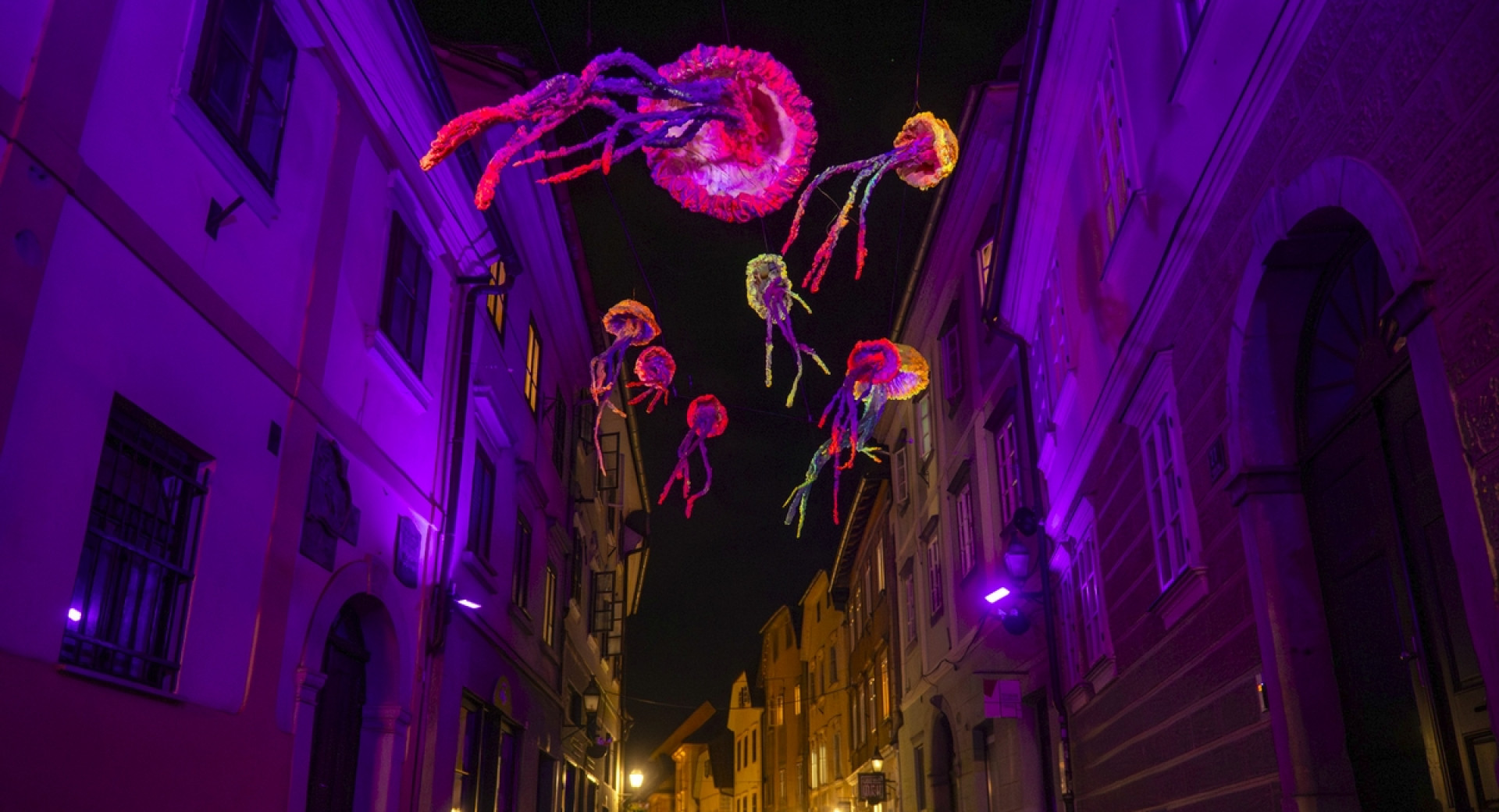 Barviti in osvetljeni liki v obliki meduz v zraku med hišami na ulici.